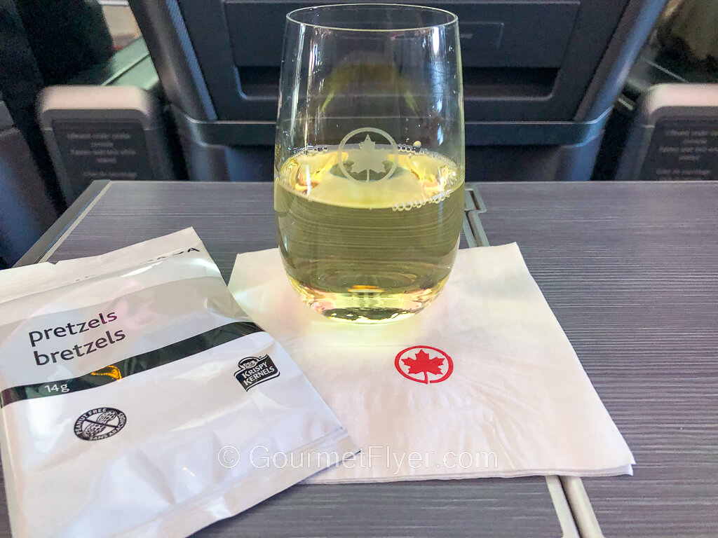 A glass of white wine placed on a napkin on top of the tray table is accompanied by a packet of pretzels.