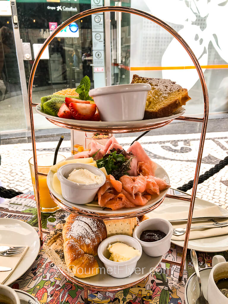 A 3-tiered serving platter contains fruits on top, ham, cheese, and salmon in the middle, and a variety of pastries on the bottom.