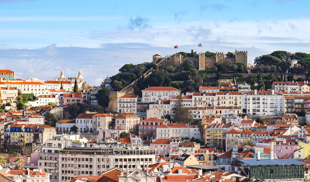 A castle stands on the top of the hill with dozens of buildings with red tile roofs below it.