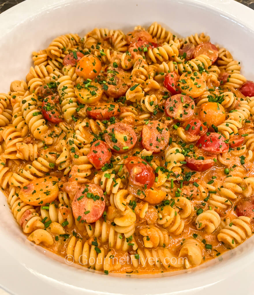 A bowl of twisty pasta is served with a red tomato-based sauce garnished with sliced cherry tomatoes and chives.