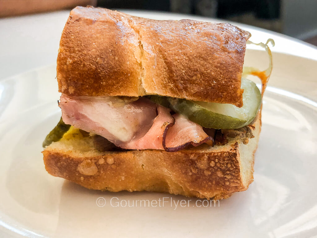 A Cuban sandwich served on a toasted roll is served on a small white plate.