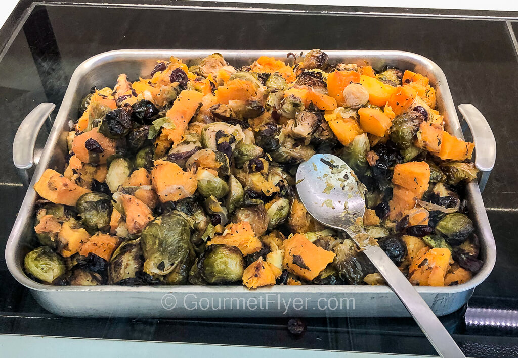 A tray of diced yellow squash and Brussel sprouts is displayed on a counter with a serving spoon.
