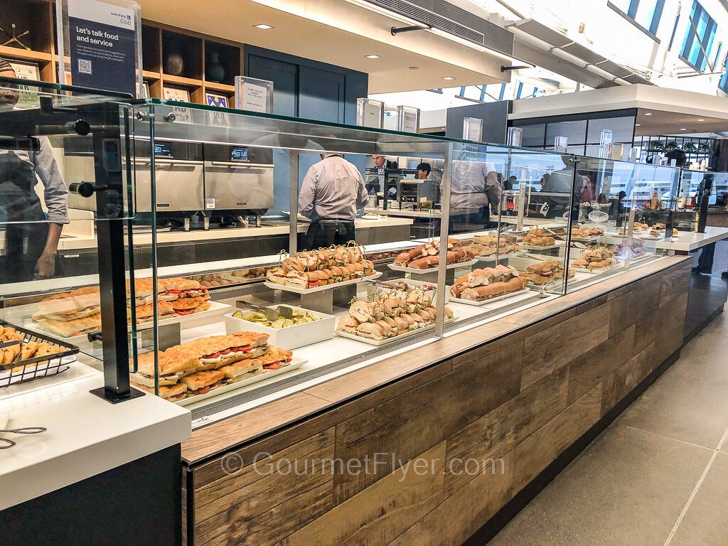 A review of the United Club at Newark EWR airport features a countertop partitioned by glass displaying many different sandwiches.