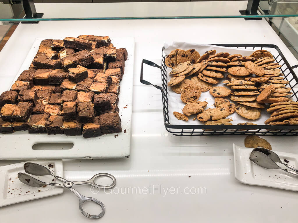 A tray of brownie lies next to a tray of chocolate chip cookies.
