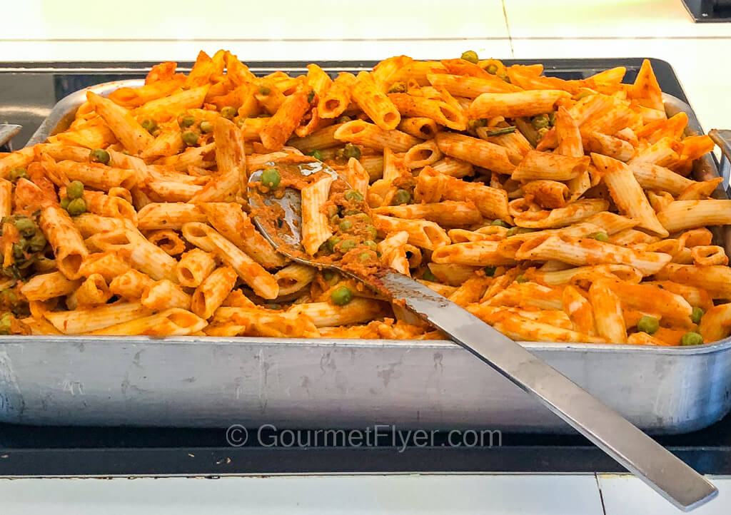 A large aluminum tray is filled with penne pasta tossed in an orange color sauce and garnished with parsleys. 