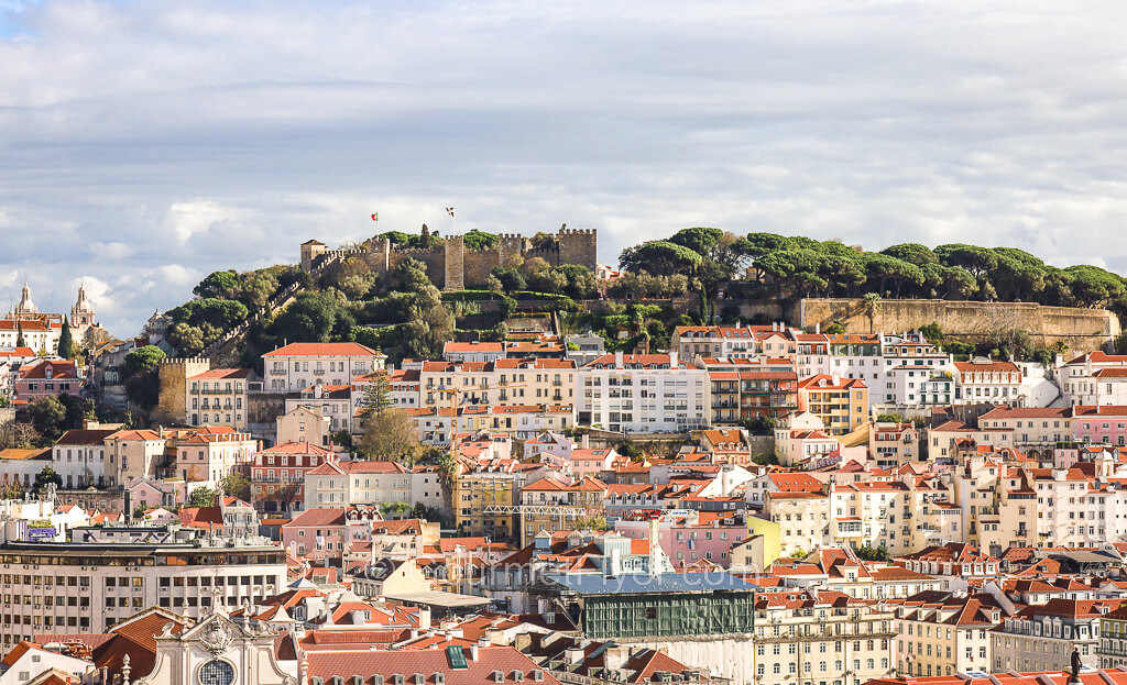 A classic scene of Lisbon's buildings with pastel color walls and red tile roofs can be seen populating a hill with a castle in the background.