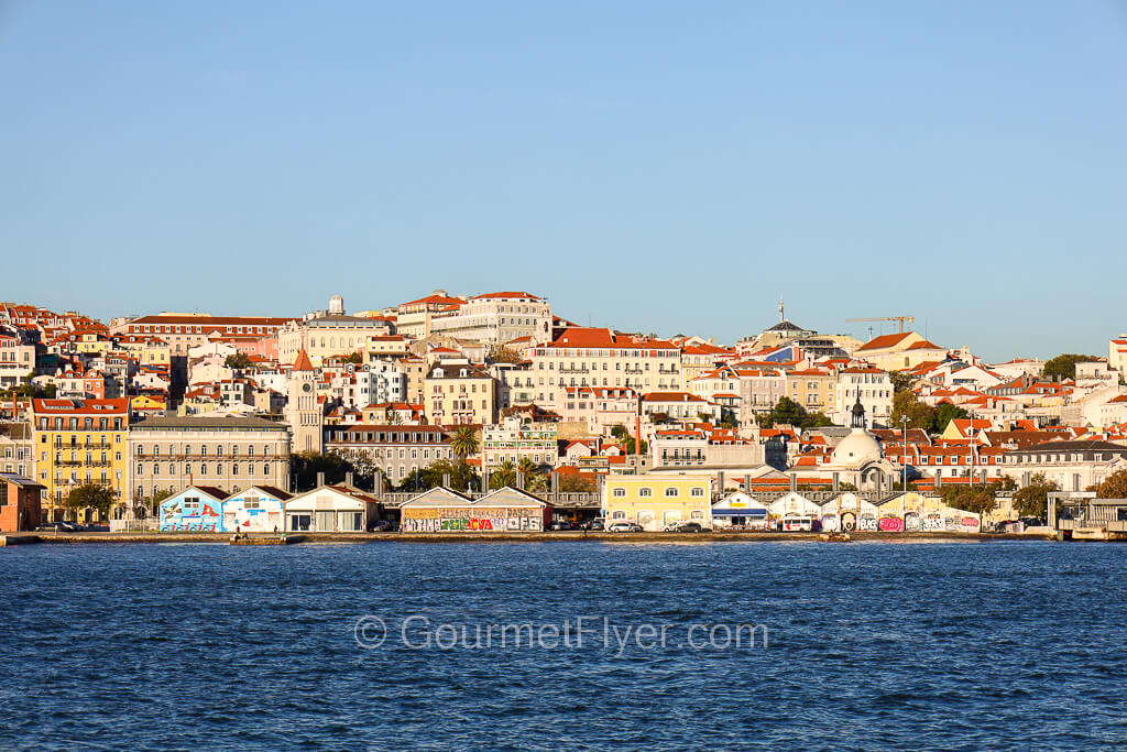 A view of Lisbon from the water shows buildings with pastel color walls and red tile roofs occupying a hill.