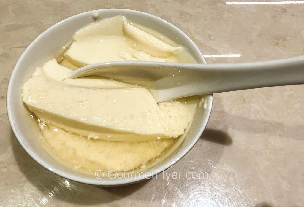 Bean curd is served with a light-yellow syrup in a small bowl with a ceramic spoon.