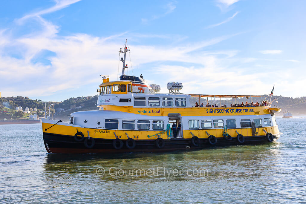 A yellow ferry sails along the water of the Tagus River under a sunny blue sky.