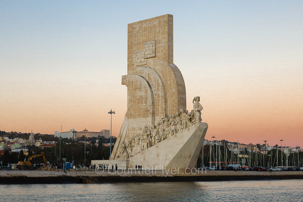 A sculpture sits on the bank of the water with a ship shaped structure carrying dozens of figures on it.