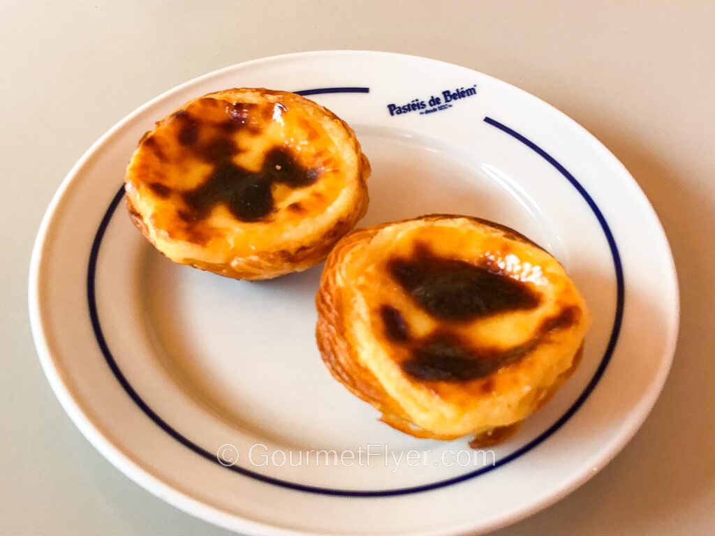 Two egg custard tarts with the top of their fillings slightly burnt are served on a small white plate.