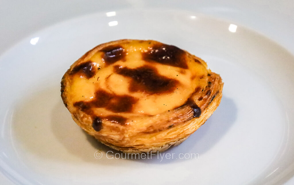 An egg custard tart with its topping charred at various spots is served on a white ceramic plate.