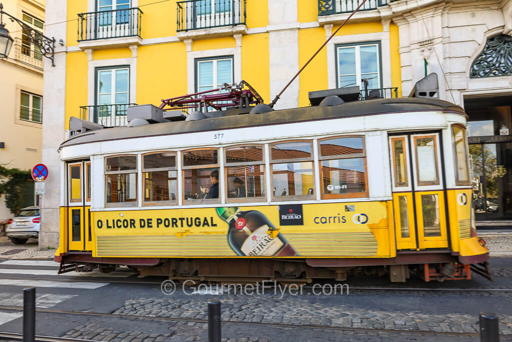 A yellow tram travels on its track on a slightly uphill road with a yellow building in the background.