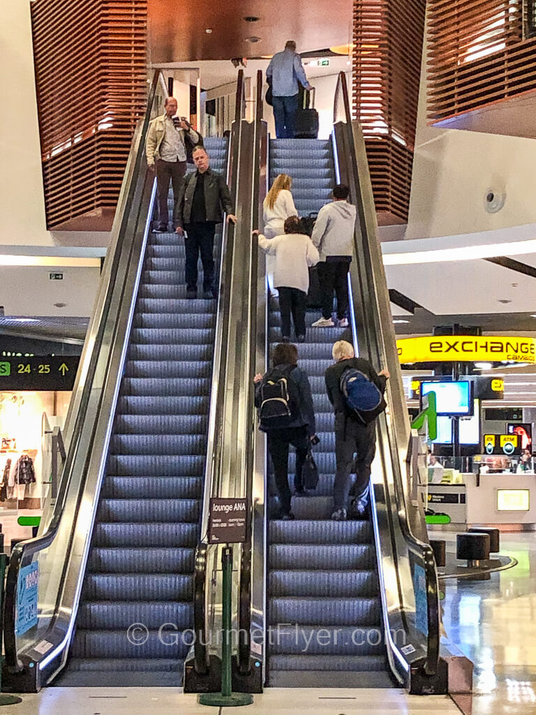 Passengers are ascending and descending on two sets of long escalators with retail stores in the background.