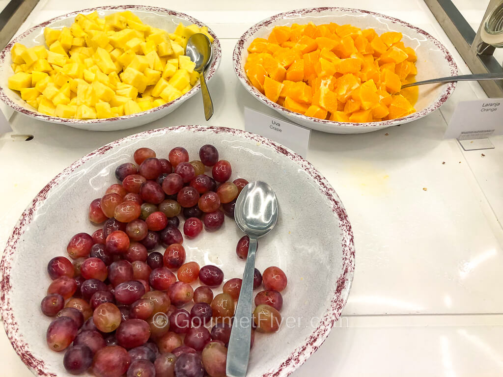 Three dishes on a countertop contain cut pineapples, pieces of peaches, and grapes.