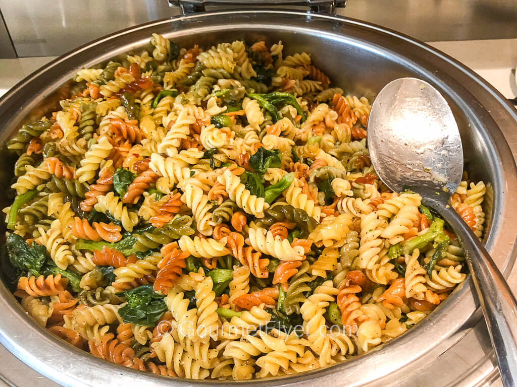 A silver serving bowl is filled with curly pasta of different colors mixed with green vegetables.