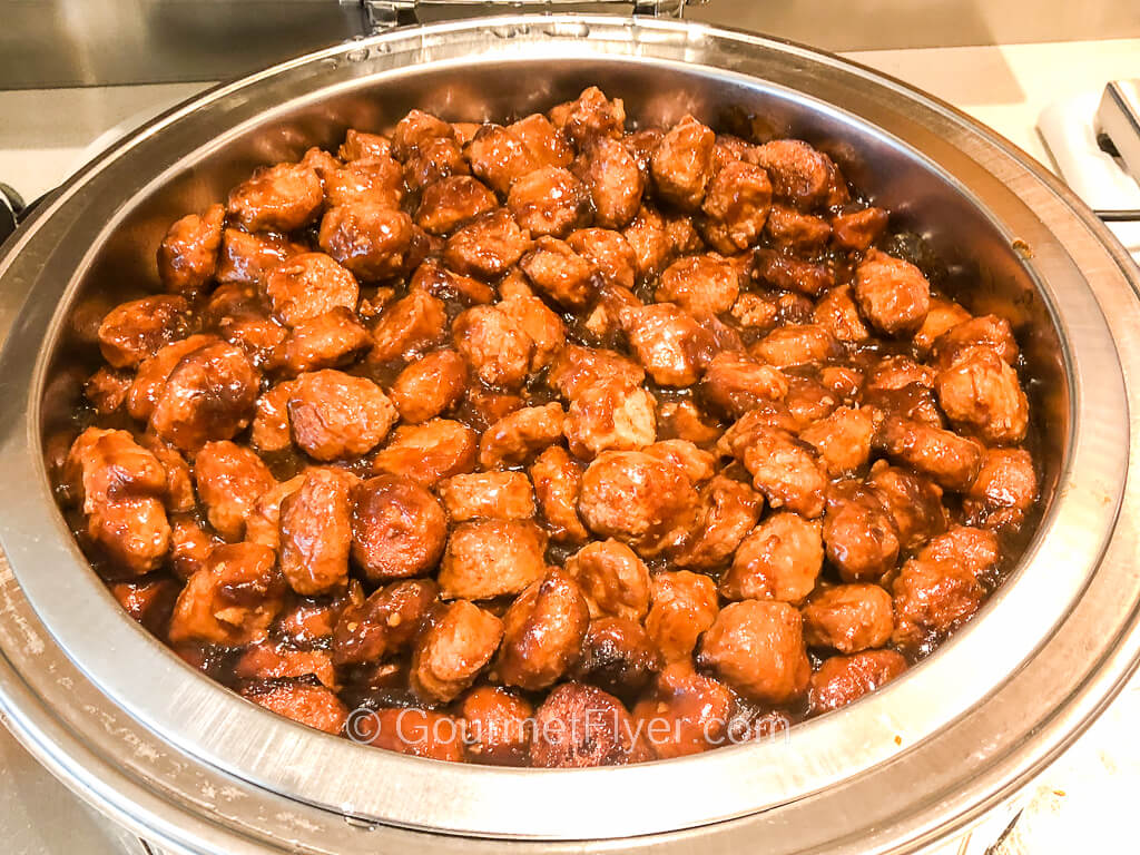A large chaffing dish is filled with cubes of chicken meat that are deep-fried and coated with a brown sauce.