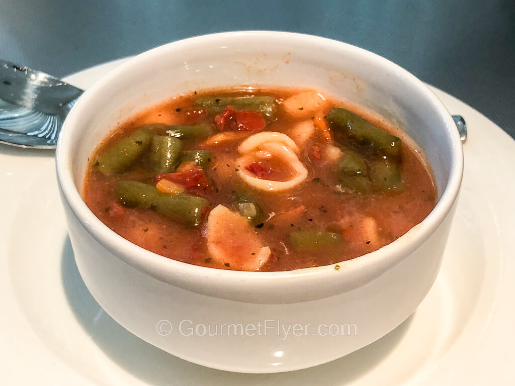 A bowl of red tomato-based soup contains green beans and pasta shells. 