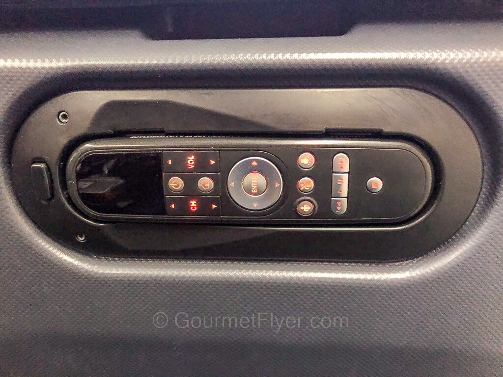 A remote control with lit orange buttons sits sideways in a bracket on the seat back entertainment console.