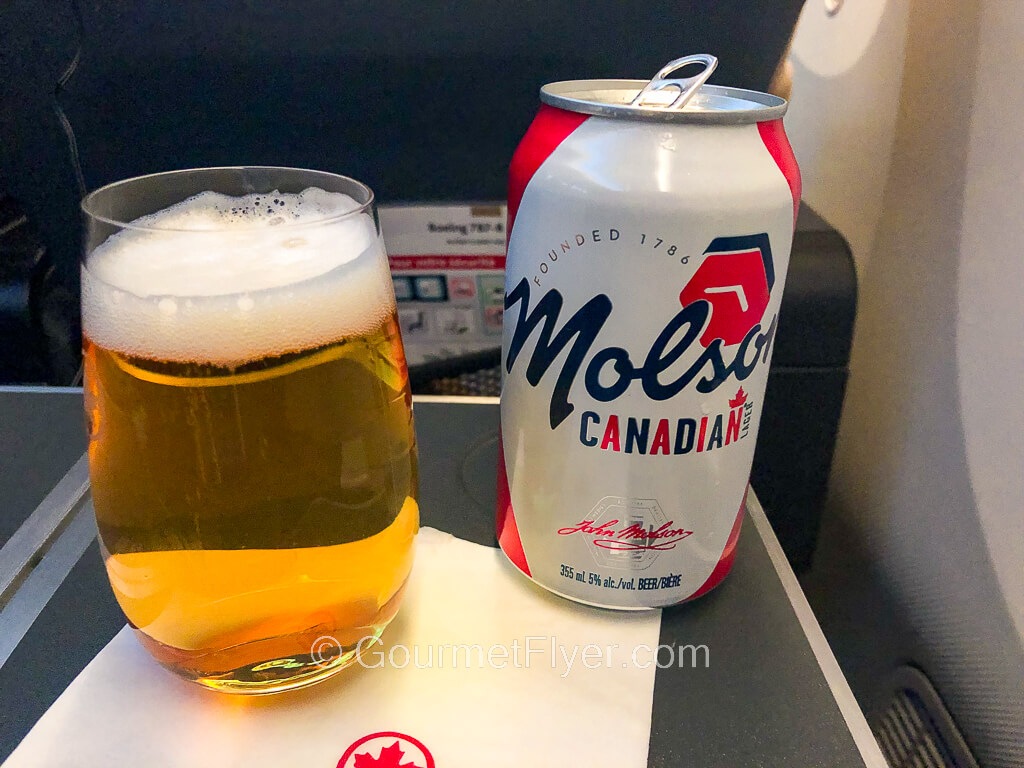 A glass full of beer is placed on a white napkin on a tray table accompanied by a Molson beer can.