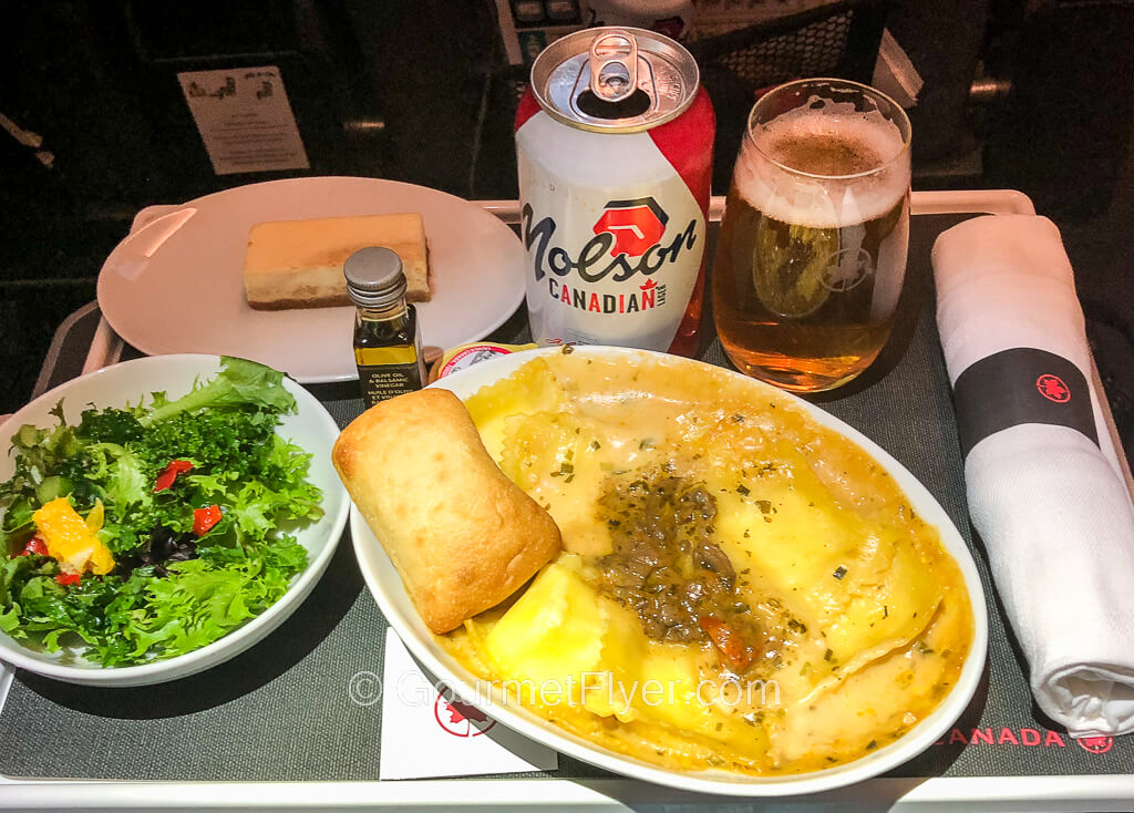 A dinner tray contains a dish of ravioli accompanied by a green salad, dessert, rolled-up silverware, and a glass of beer.
