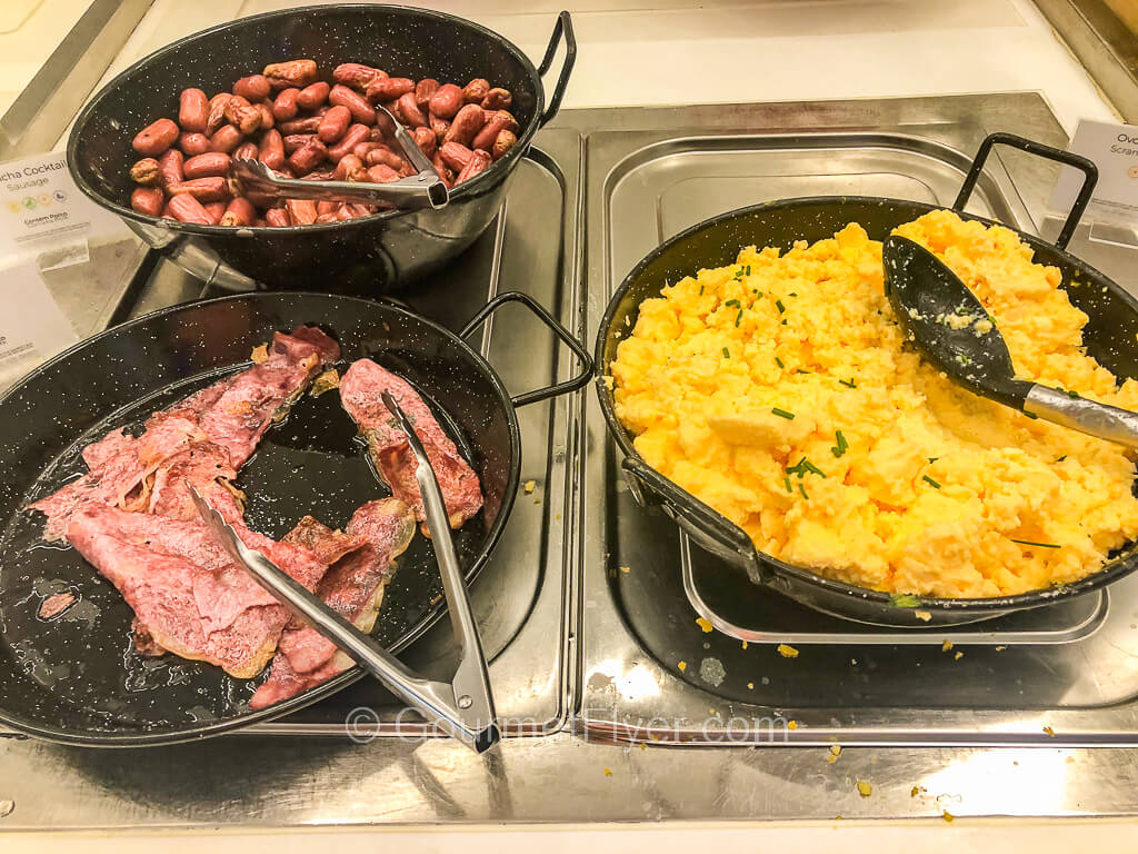 Three metal hot platters are filled with scrambled eggs. sausages, and slices of grilled bacon.