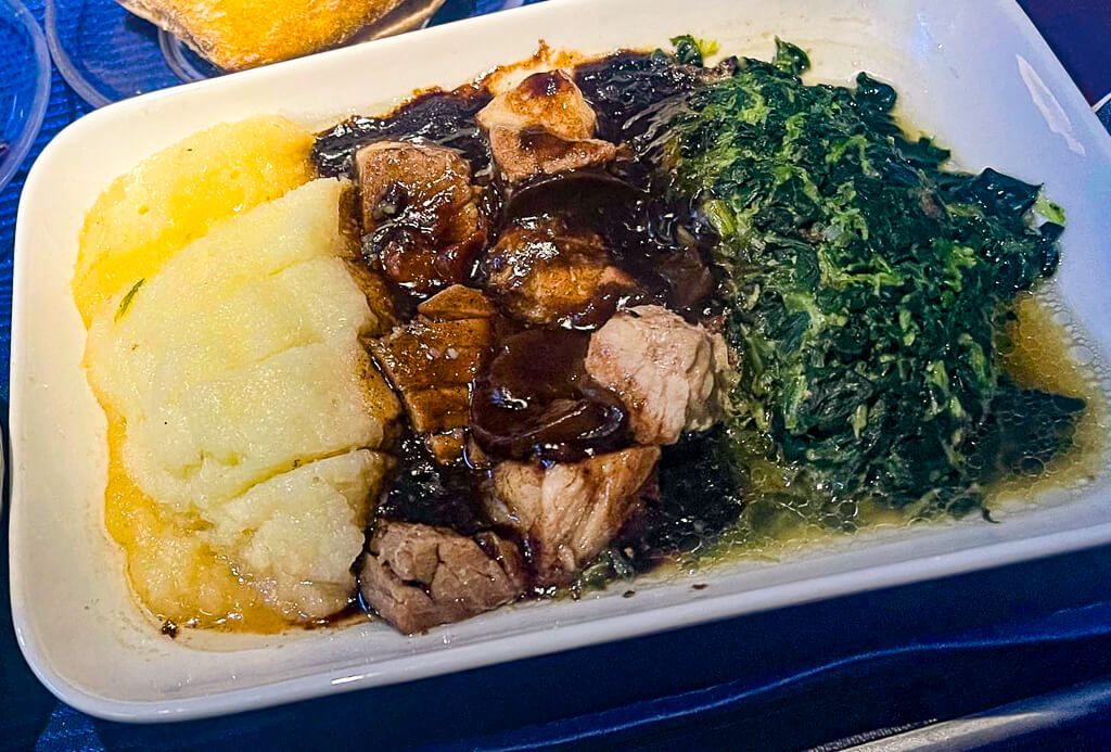 A dish of cubed meat in a dark barbeque sauce is accompanied by mashed potato on its left and spinach on its right.
