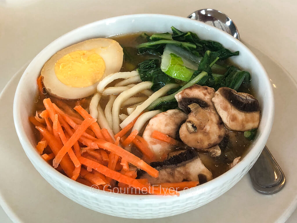 A bowl of soup noodle is topped with shredded carrots, vegetables, and half a hard-boiled egg.