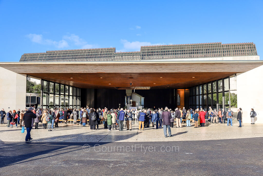 Many people are attending a liturgy in the open space of a chapel under sunny blue skies.