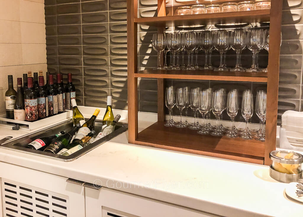 Many bottles of red and white wines are accompanied by a shelf of wine glasses and flutes.