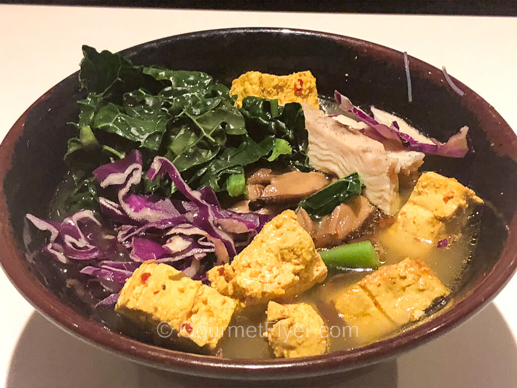A bowl of soup noodles topped with chicken, tofu, green vegetables, and red cabbage.