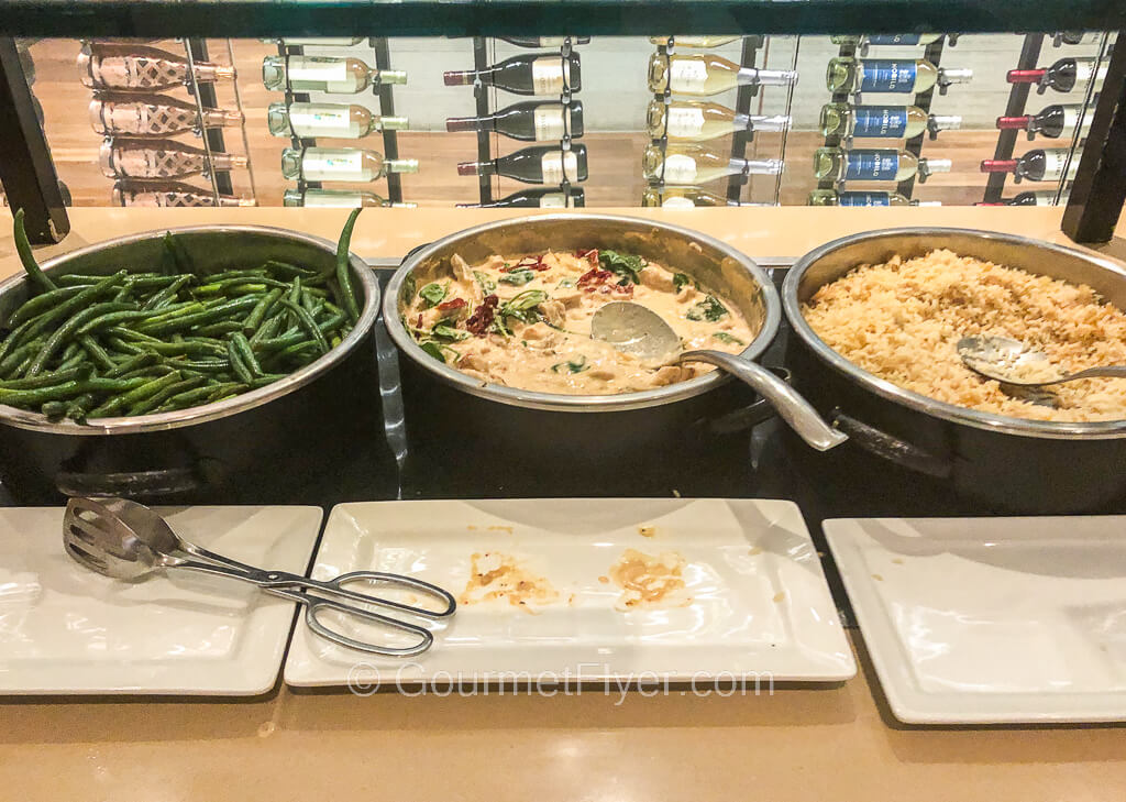 Chaffing bowls containing green vegetables, creamy chicken and rice pilaf are placed on a buffet counter.