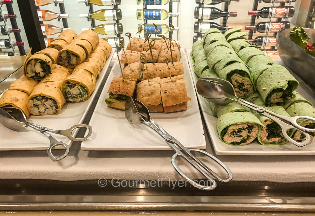 Three long vertical trays side by side contain chicken wraps, a cutup sub sandwich, and green tuna wraps.