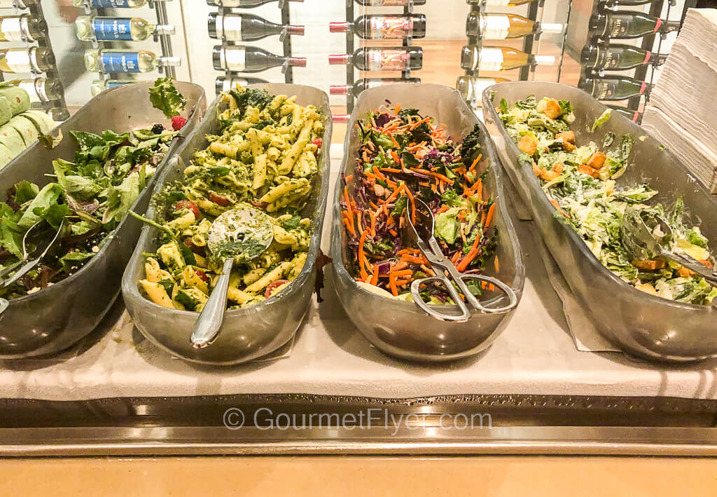 Four long vertical serving bowls containing different salads are placed side by side on a countertop.