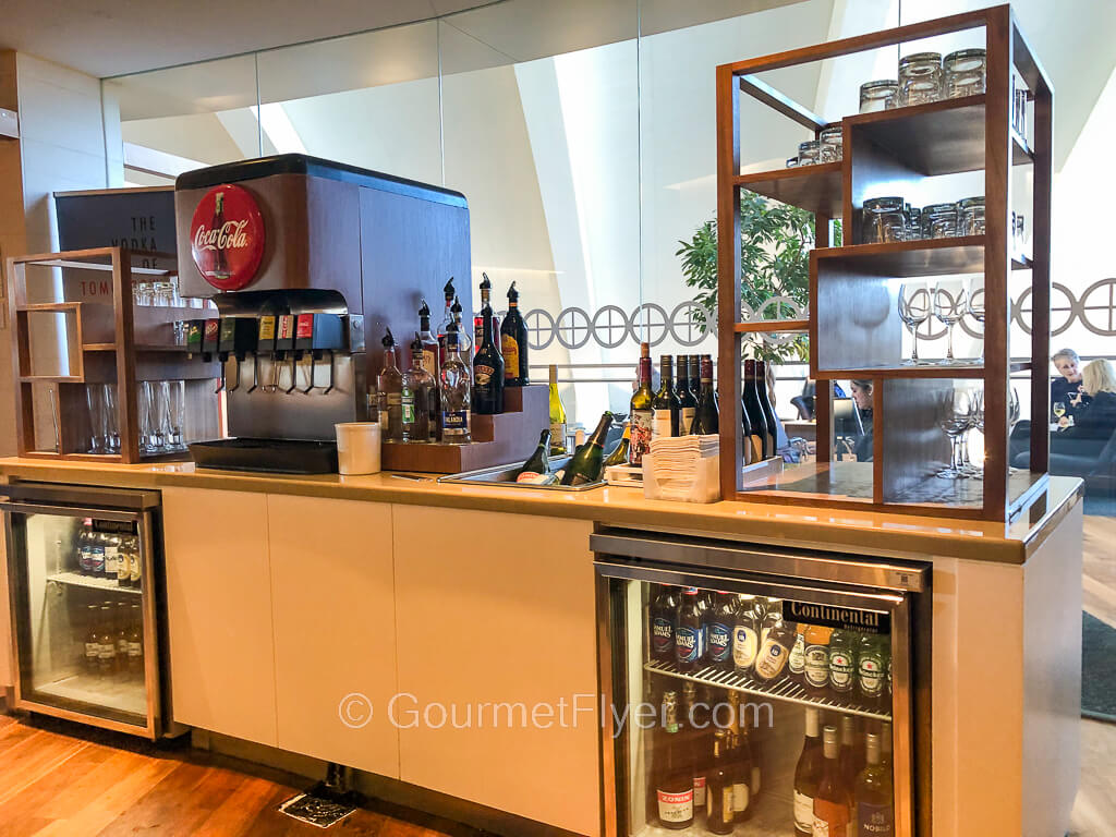 A long counter with a coke machine and bottles of liqueurs and wines and top and two mini fridges with glass doors in the bottom. 
