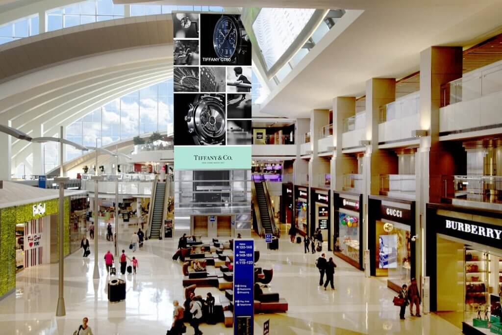 At the lobby of an airport terminal is a tall floor to ceiling time tower with computer generated images and a pair of elevators at its bottom.