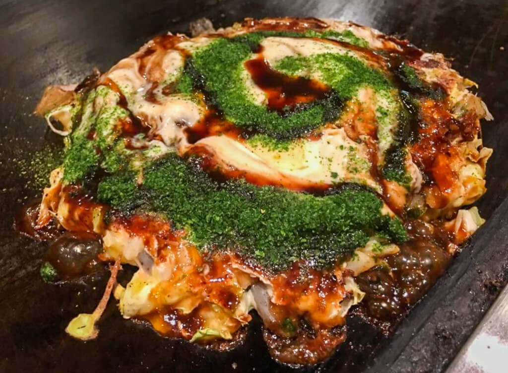 A grilled pancake like dish that is topped with a brown sauce and green powders.