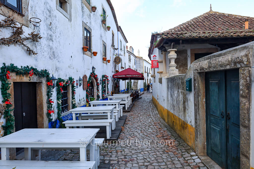 A narrow quaint cobblestone street is lined with white dining tables and benches from a restaurant on its left.