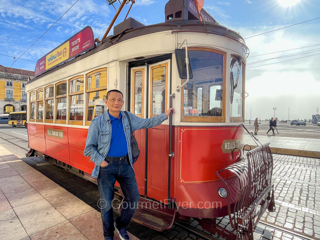 A man is posing in front of a tourist red tram, holding the handrail outside its front door.