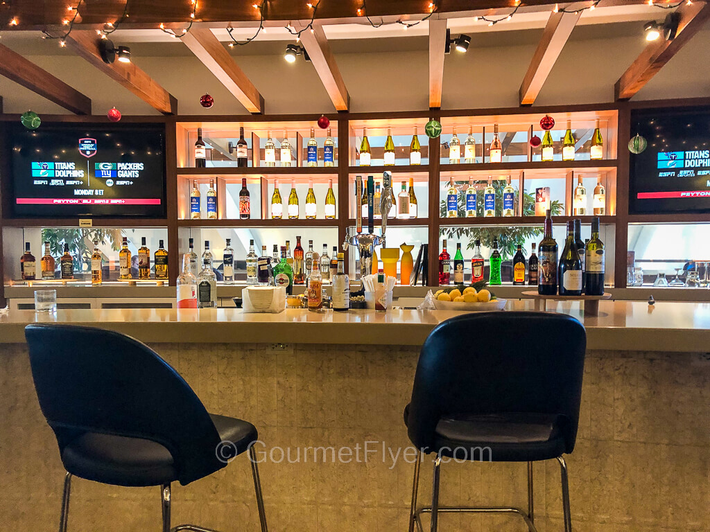 A bar has large and brightly lit shelves of bottles behind it and is accompanied by two TVs.