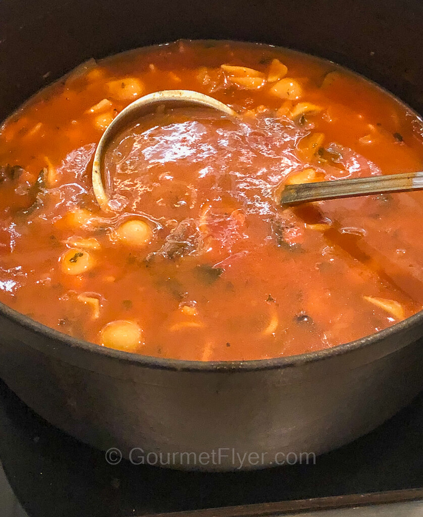 A serving bowl is filled with a red tomato-based soup loaded with pasta shells and diced vegetables.