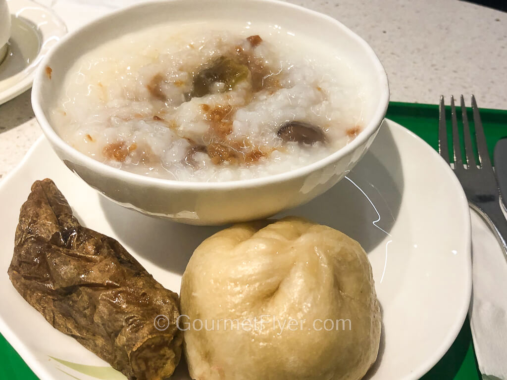 A plate contains a bowl of congee with toppings together with a steamed bun and a piece of yam.