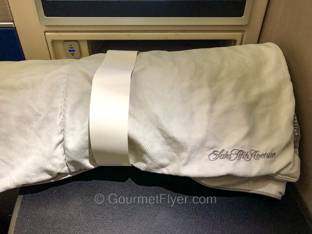 A light grey color duvet is rolled up and placed on the tray table in front of the seat.