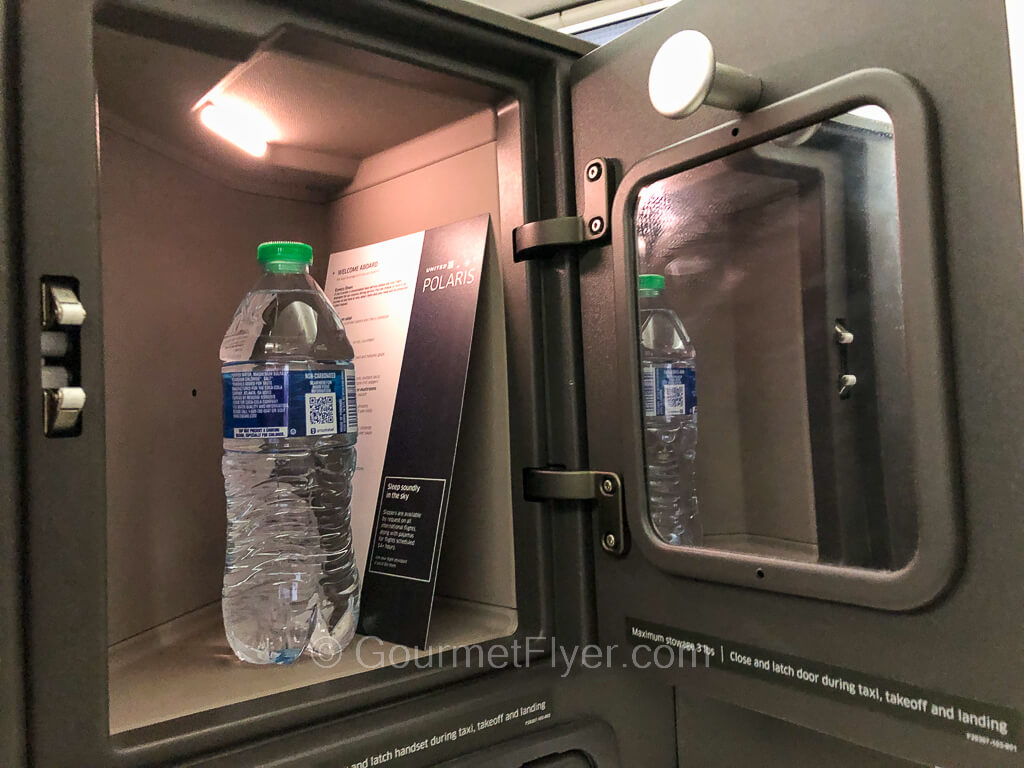 An opened small storage compartment at a business class seat has a bottle of water in it. along with a mirror on the door.
