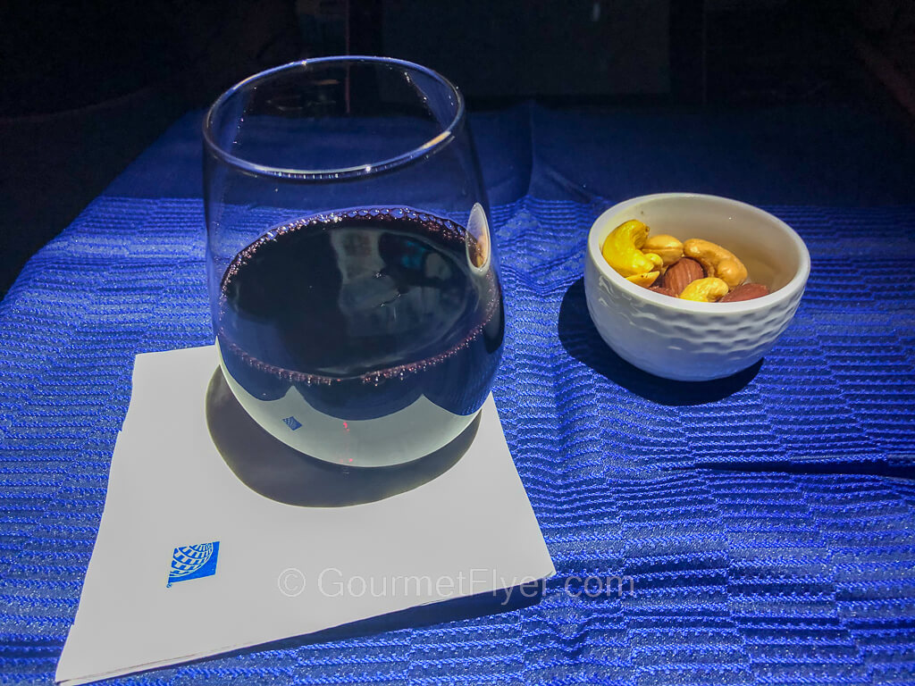 A glass of red wine sits on a table with a blue tablecloth and accompanied by a small serving of nuts.