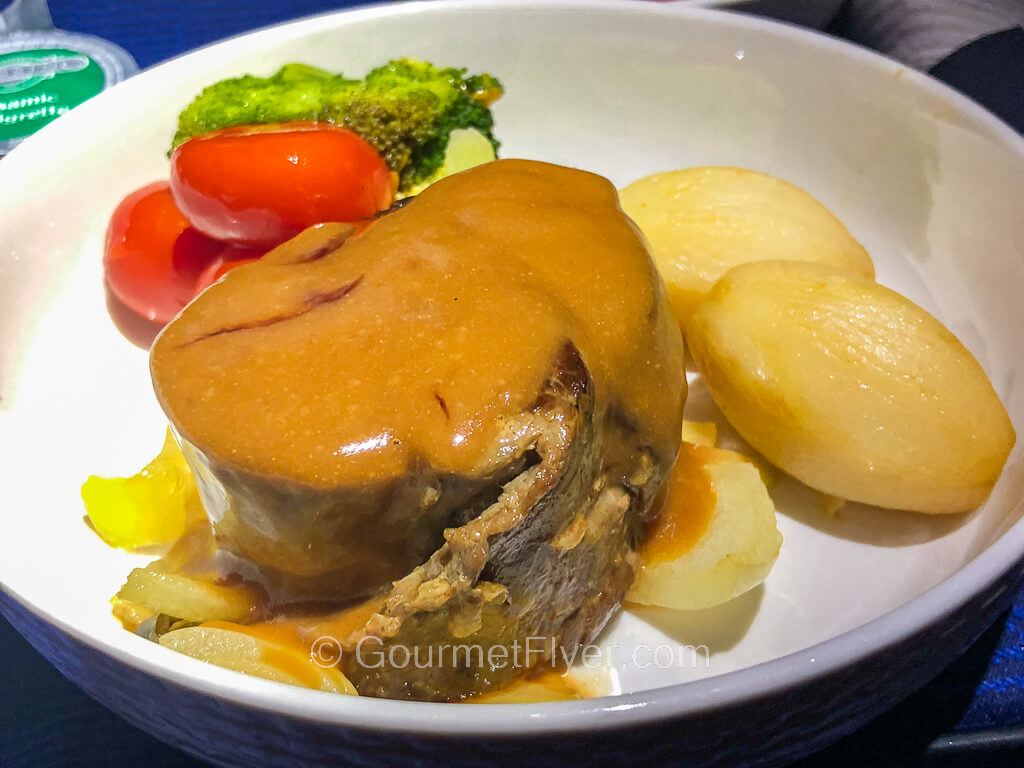 A rolled up flat iron steak topped with brown gravy is accompanied by potatoes and vegetables.