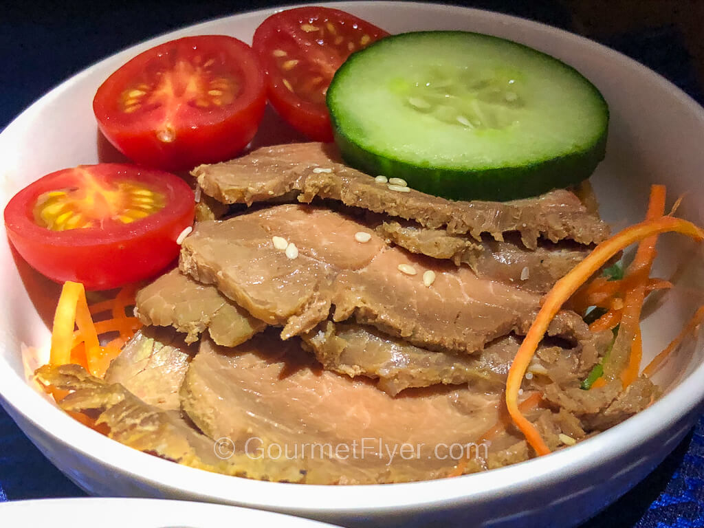 Sliced beef in a bowl is garnished with cherry tomato halves and a slice of cucumber.