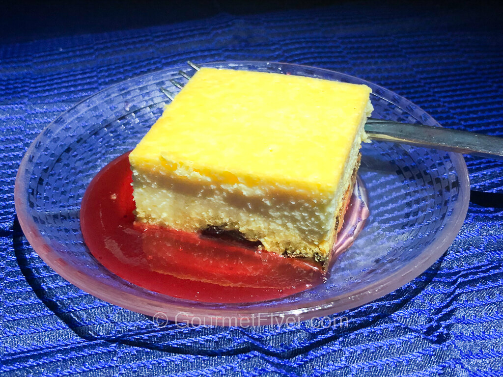 A square slice of cheesecake with a yellow top is served with red raspberry sauce.