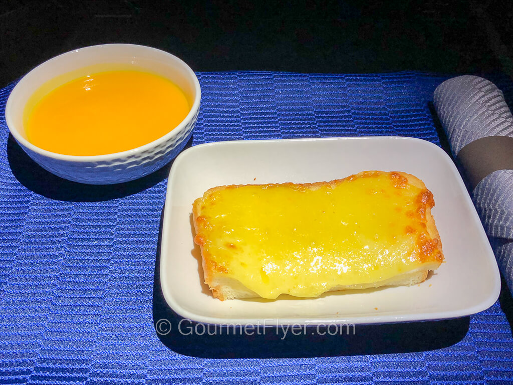 A dinner tray with blue tablecloth contains a dish with a cheese toast and a bowl of an orange color soup.