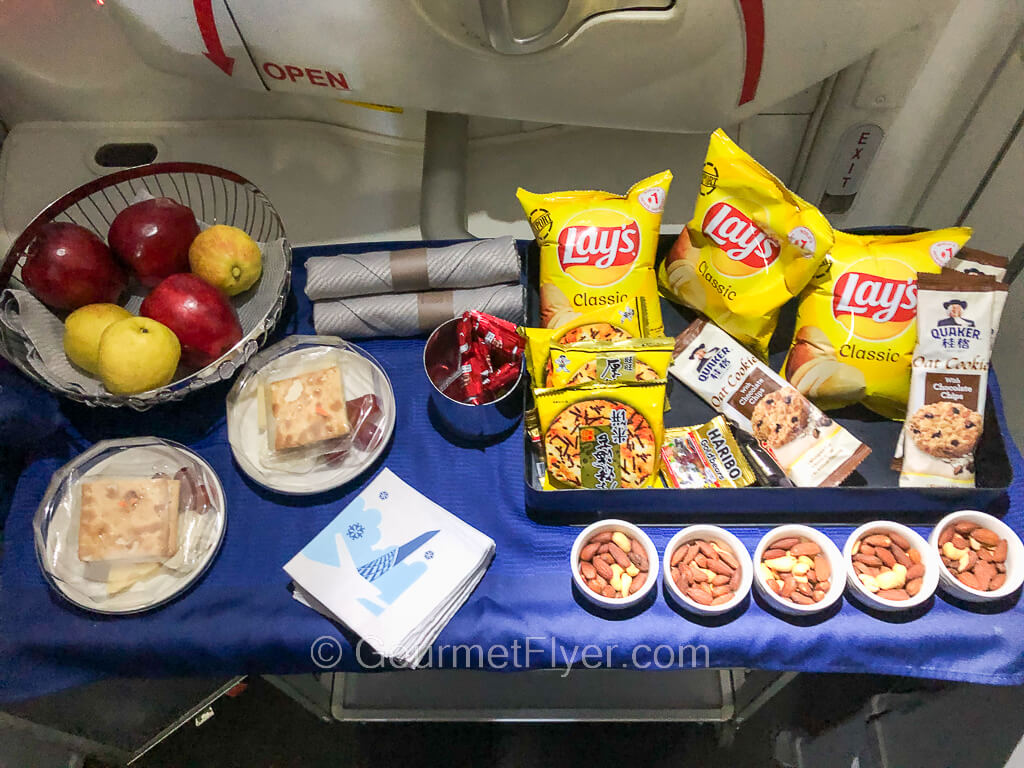 A cart with a blue tablecloth has many different food items placed on it, including a basket of fruits, bags of chips, mixed nuts, among other things.